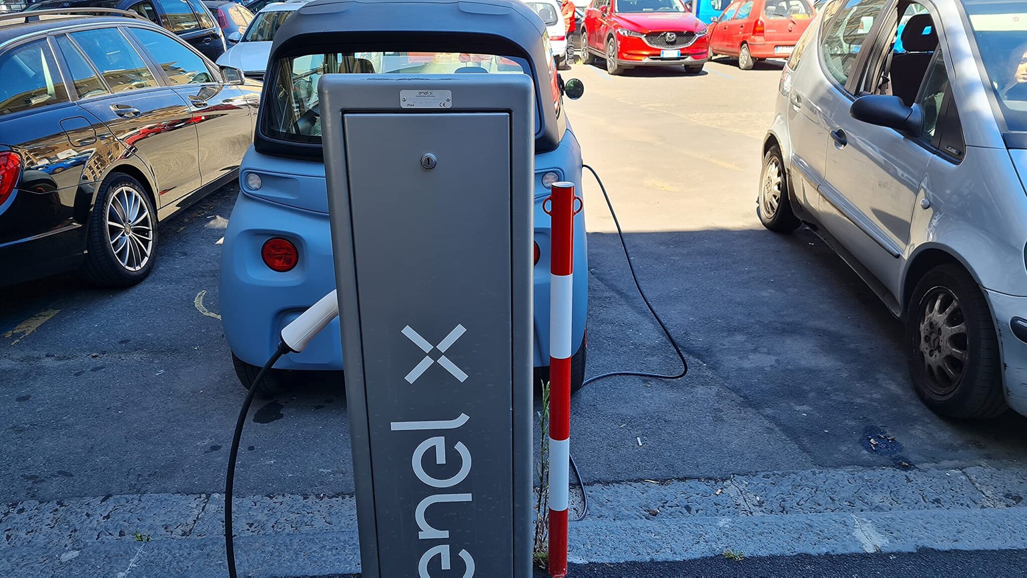 Une petite voiture électrique bleue compacte (Citroën Ami) en train de se recharger à une borne de recharge grise Enel X dans un parking urbain.