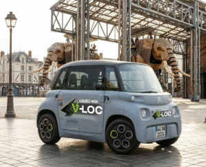 Une Citroën Ami grise de location V-LOC garée sur l'esplanade des Machines de l'Île à Nantes, avec le Grand Éléphant en arrière-plan sous les nefs.