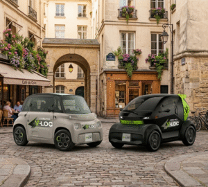 Une Citroën Ami grise et une Mobilize Duo noire, toutes deux floquées du logo "V-LOC", garées côte à côte dans une rue pavée typique de Paris devant un café fleuri.