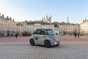 Une petite voiture électrique Citroën Ami grise avec des autocollants "V-LOC" garée au centre de la vaste Place Bellecour à Lyon, avec la colline de Fourvière et sa basilique en arrière-plan.