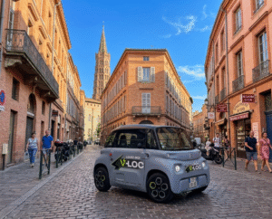 Une voiture électrique Citroën Ami grise avec le logo "V-LOC" circulant dans une rue pavée typique de Toulouse, entourée de bâtiments en briques roses sous un ciel bleu avec le clocher de la basilique Saint-Sernin en arrière-plan.